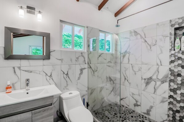 A bathroom with a white sink, a toilet, and a shower area with a glass door. There are windows above the shower, and a mirror above the sink.