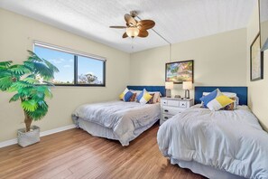 A bedroom with two beds, a ceiling fan, a window, a plant, and a painting on the wall. - Pet-friendly Condo Near the Beach w/ Boat Slip (Treasure Island)