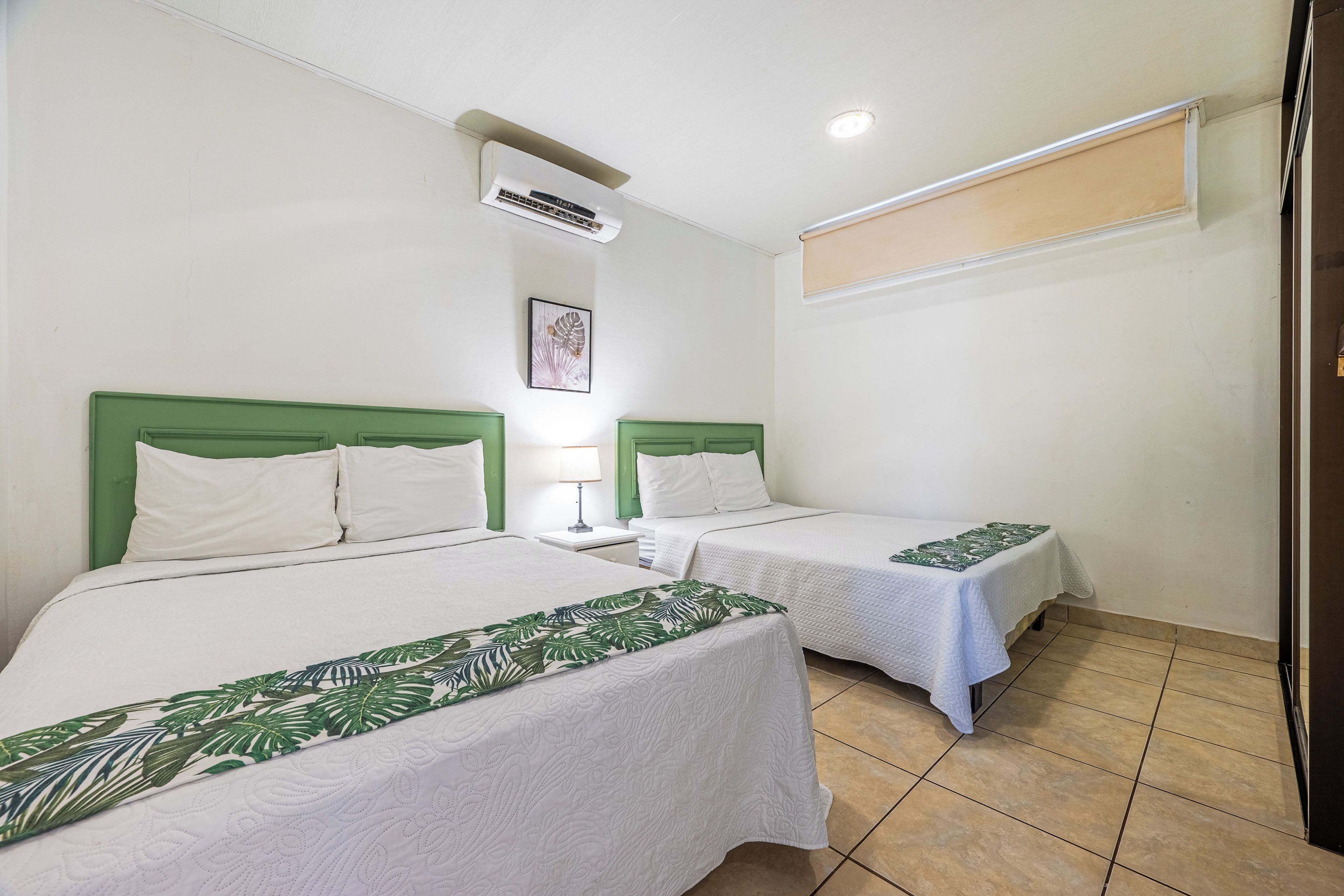 A hotel room with two beds, each with a green headboard and white bedding. There is a single bedside table with a lamp, and a ceiling-mounted air conditioning unit.