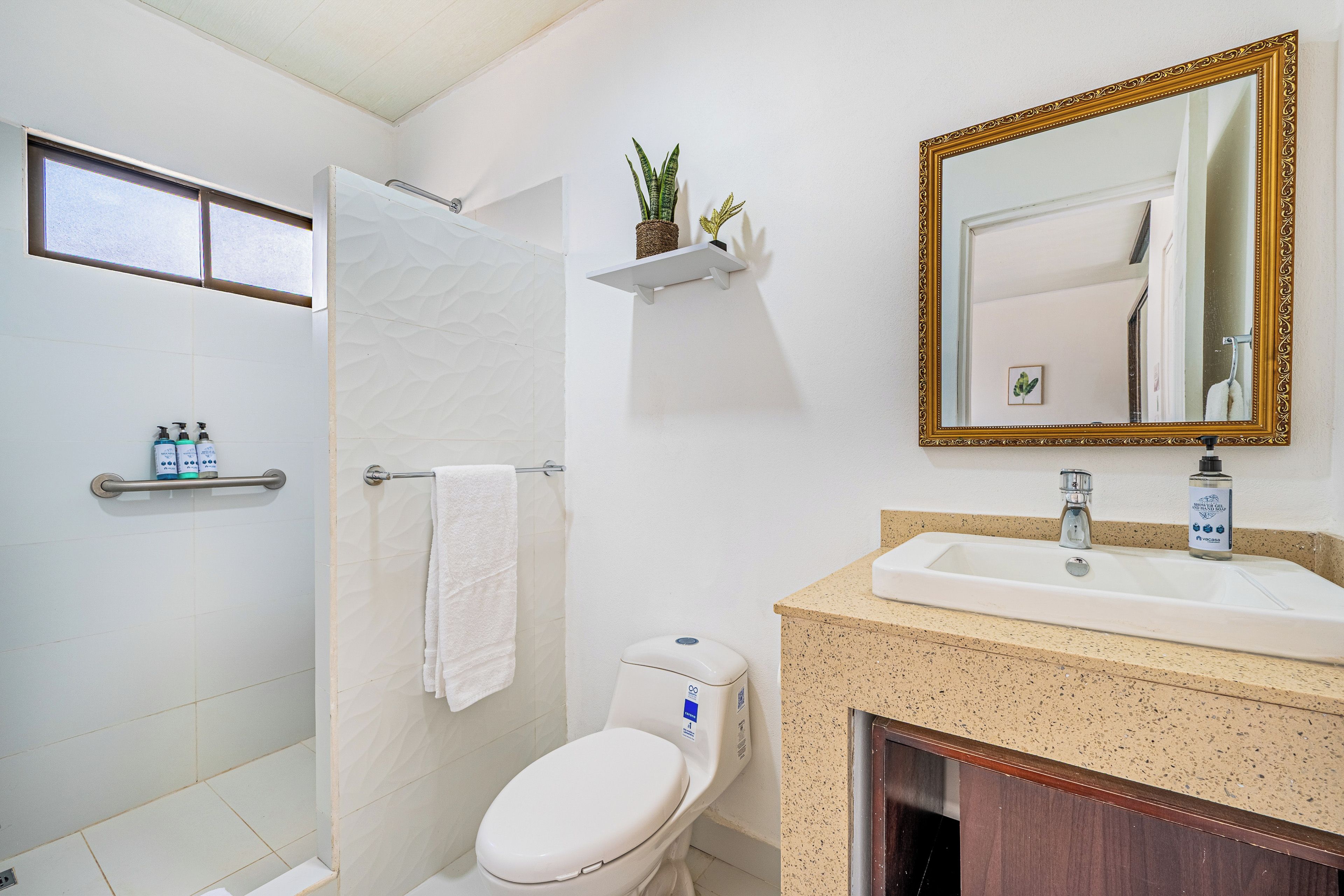 Ein Badezimmer mit Toilette, Waschbecken und Dusche. Handtuchhalter, Ablage mit Pflegeprodukten und ein Spiegel mit aufwendigem Rahmen sind vorhanden.
