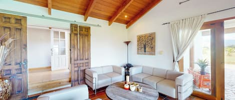 A spacious living area with a high wooden ceiling, a large white sofa set, a round coffee table, and a view through a glass door.