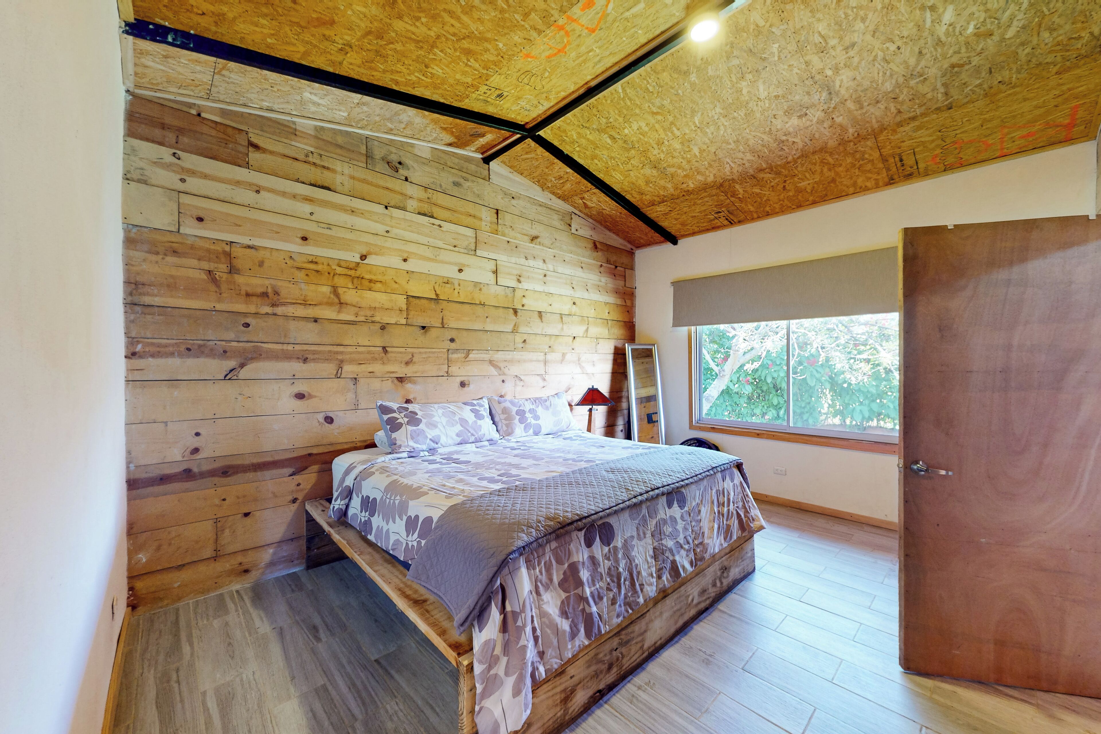 A bedroom with a wooden slat ceiling, a bed with patterned bedding, a window with a view of greenery, and a door.