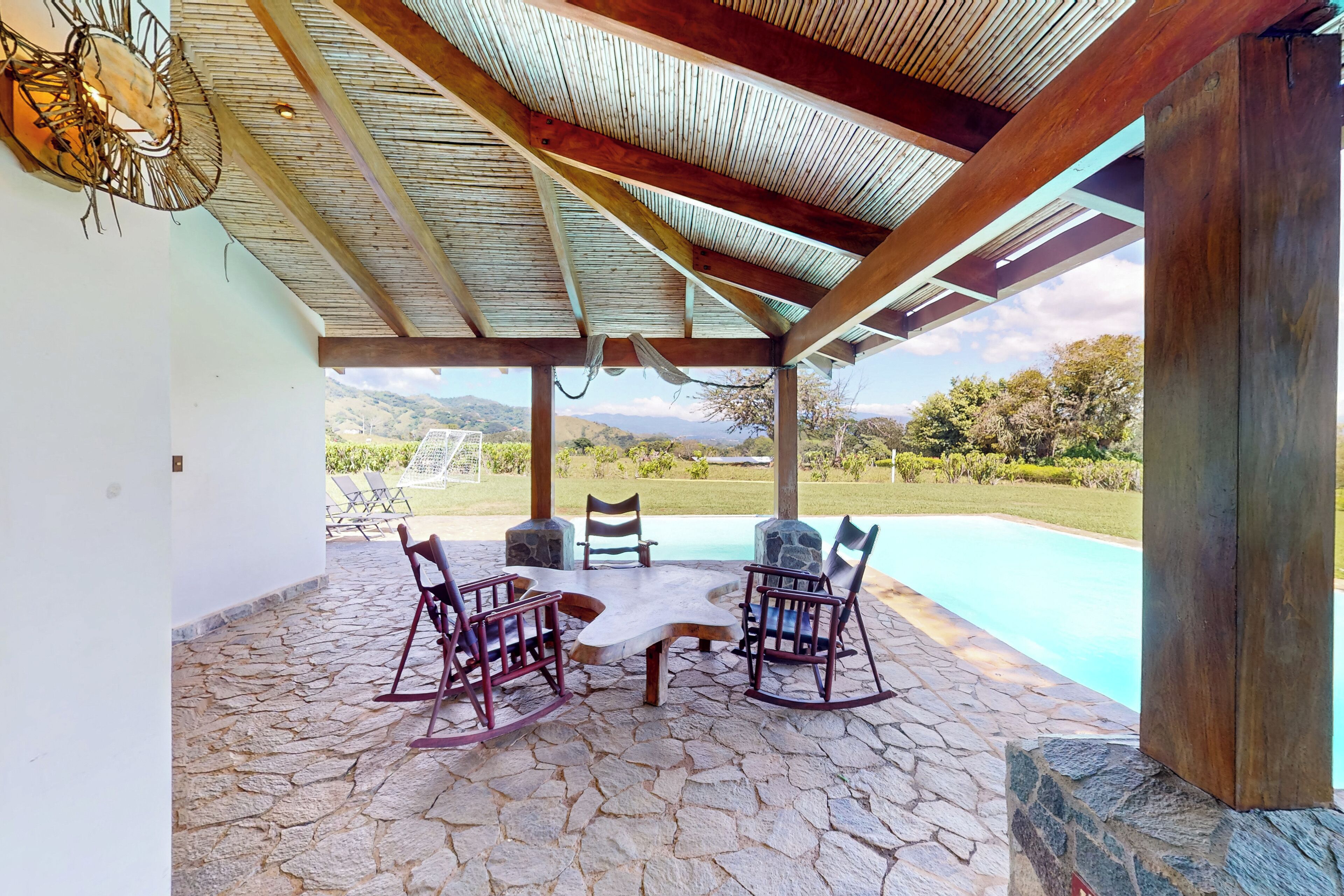 A covered outdoor seating area with a stone floor, a table, and four rocking chairs.