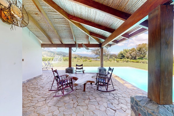 A covered outdoor seating area with a stone floor, a table, and four rocking chairs.