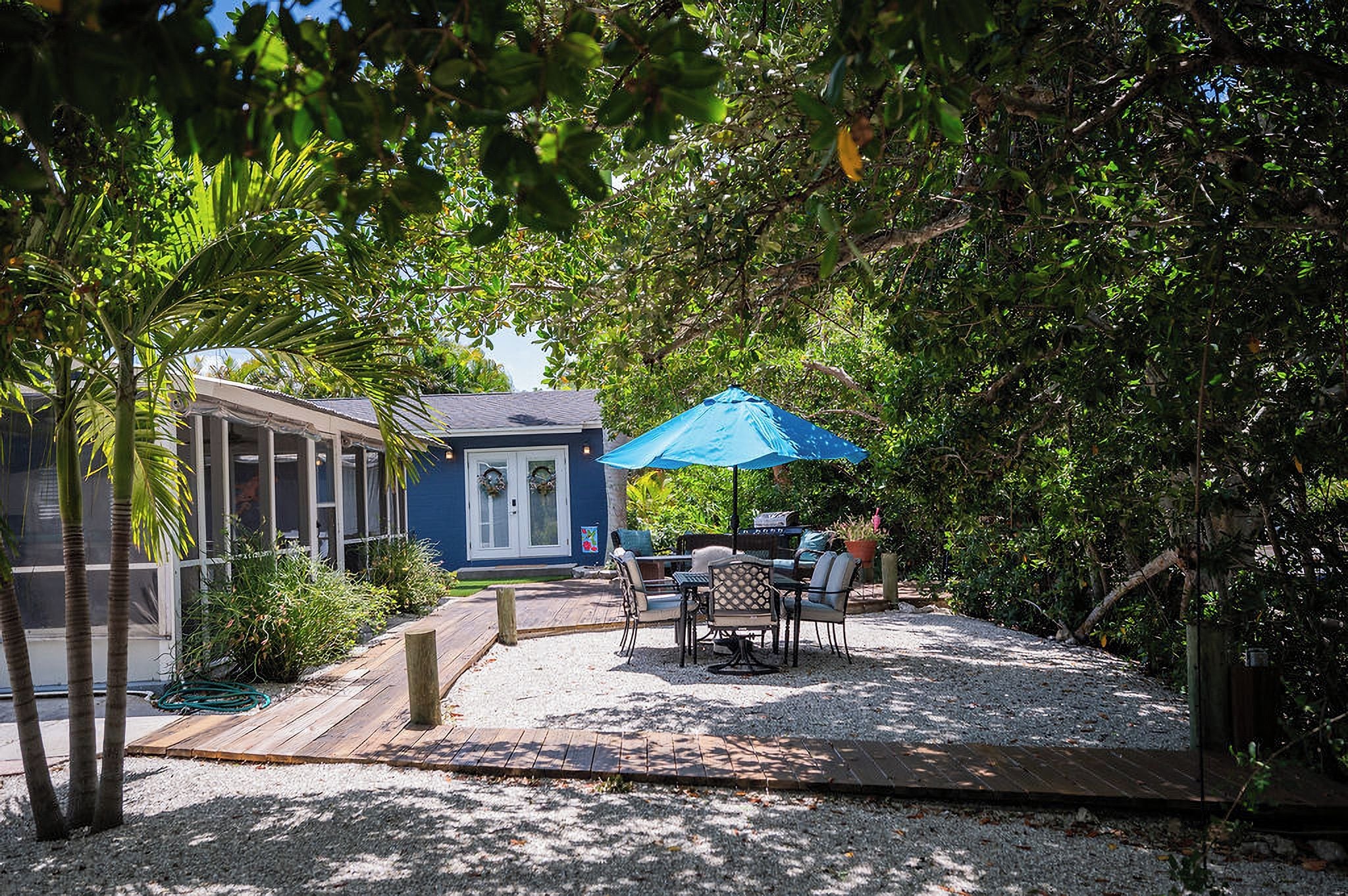 A covered outdoor seating area with a table and chairs, a blue umbrella, and a small building with a door and windows.