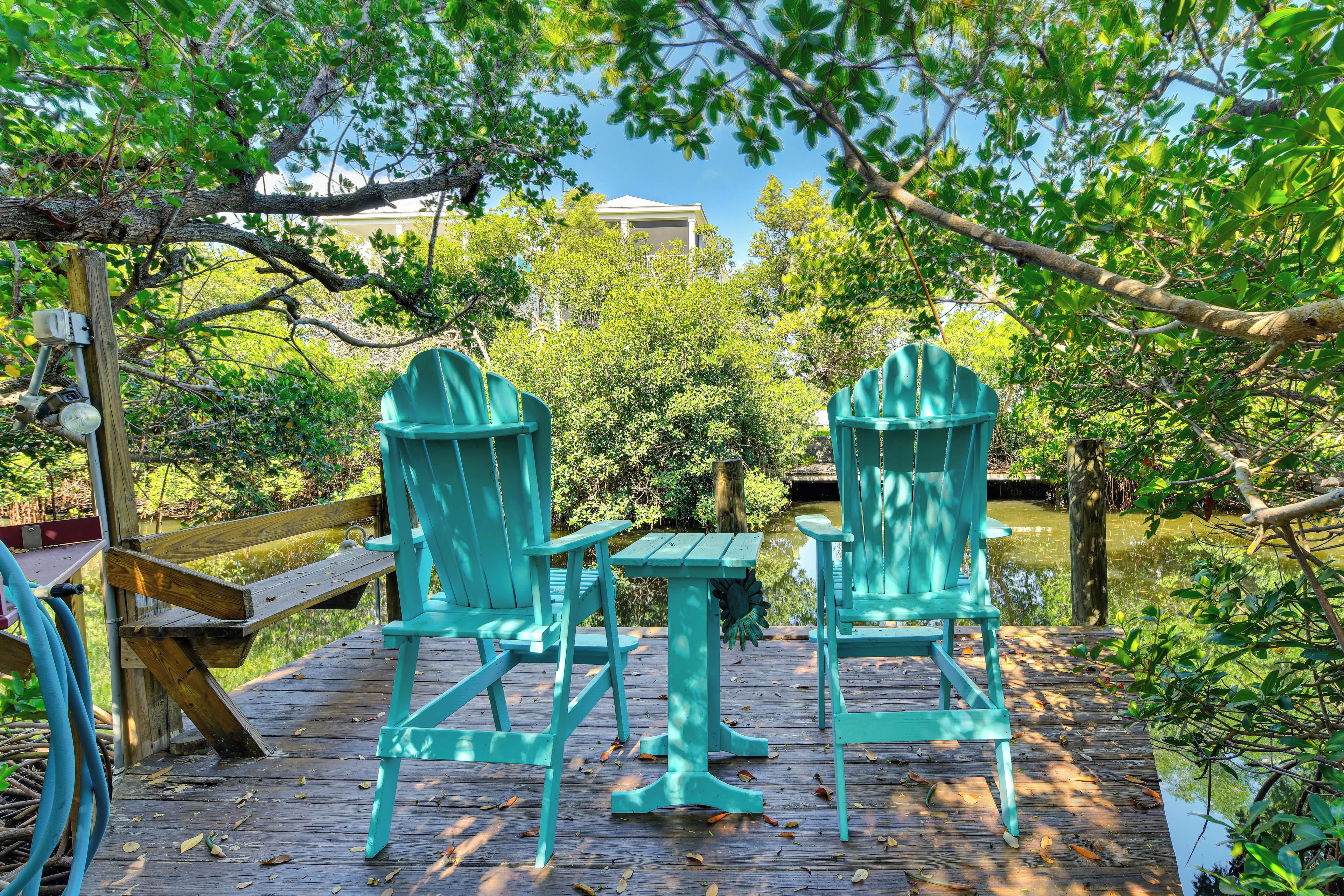 A wooden deck with two turquoise chairs and a small table, surrounded by lush greenery and a body of water.
