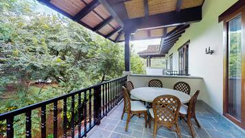 A covered balcony with a table and chairs, surrounded by a railing, overlooking a garden.