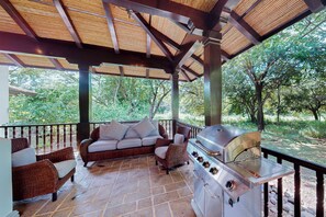 Une terrasse extérieure couverte, équipée d’un barbecue, de meubles en osier et offrant une vue sur la verdure environnante.