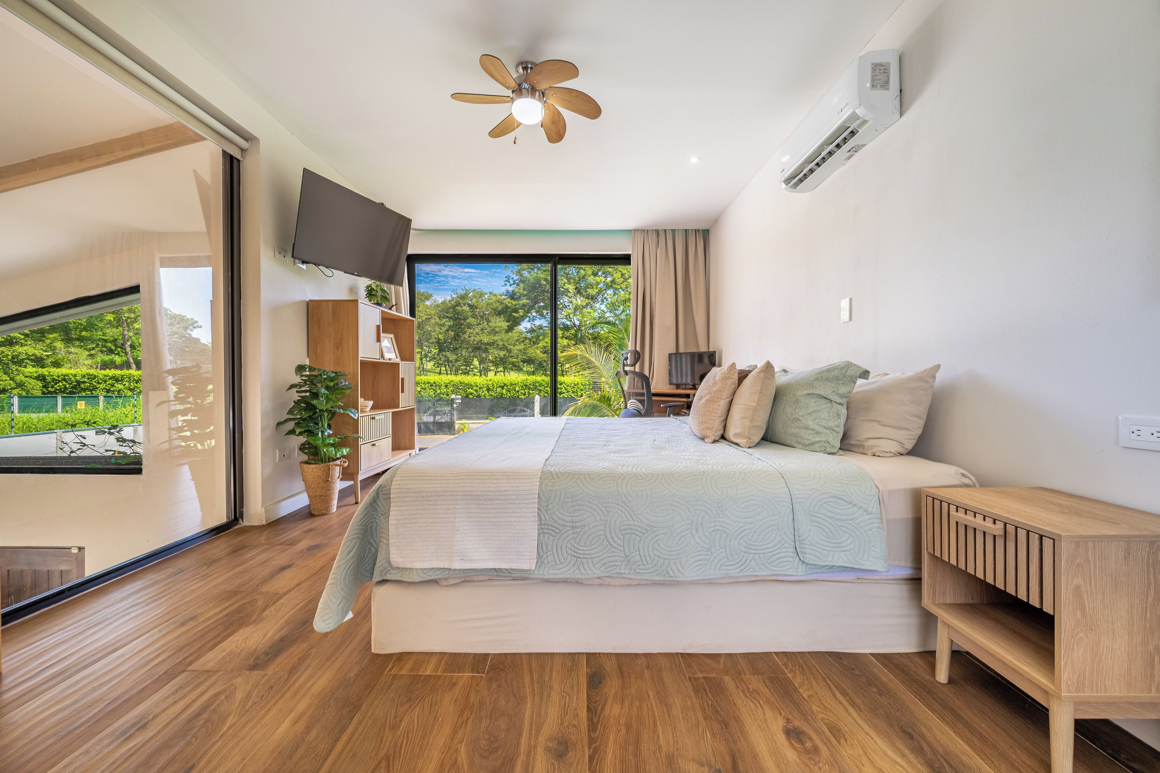 Ein Schlafzimmer mit einem Bett, einem Fernseher, einem Deckenventilator und Blick auf Grünfläche.