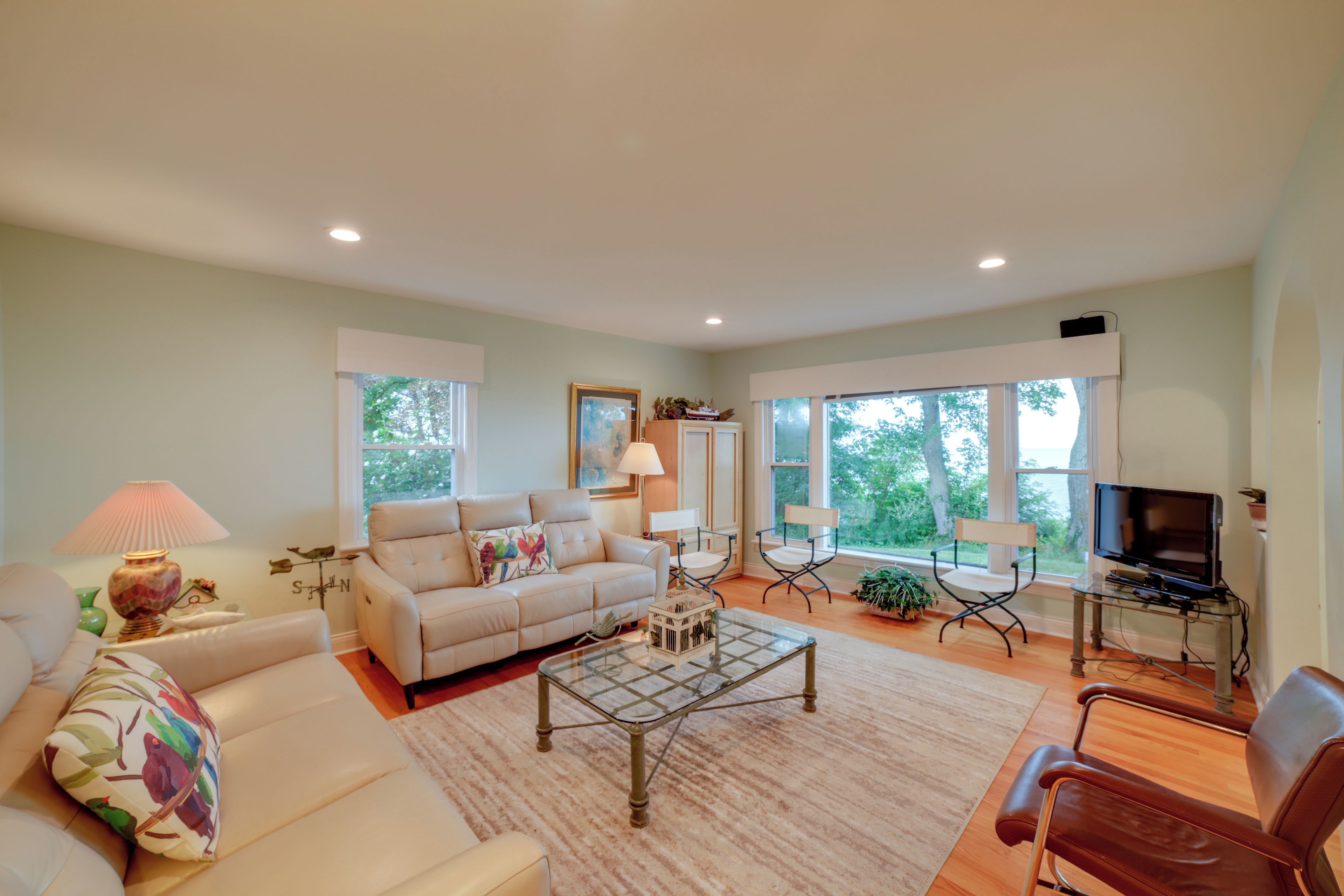 A living room with a sofa, coffee table, and chairs, a television, and large windows with a view of trees.