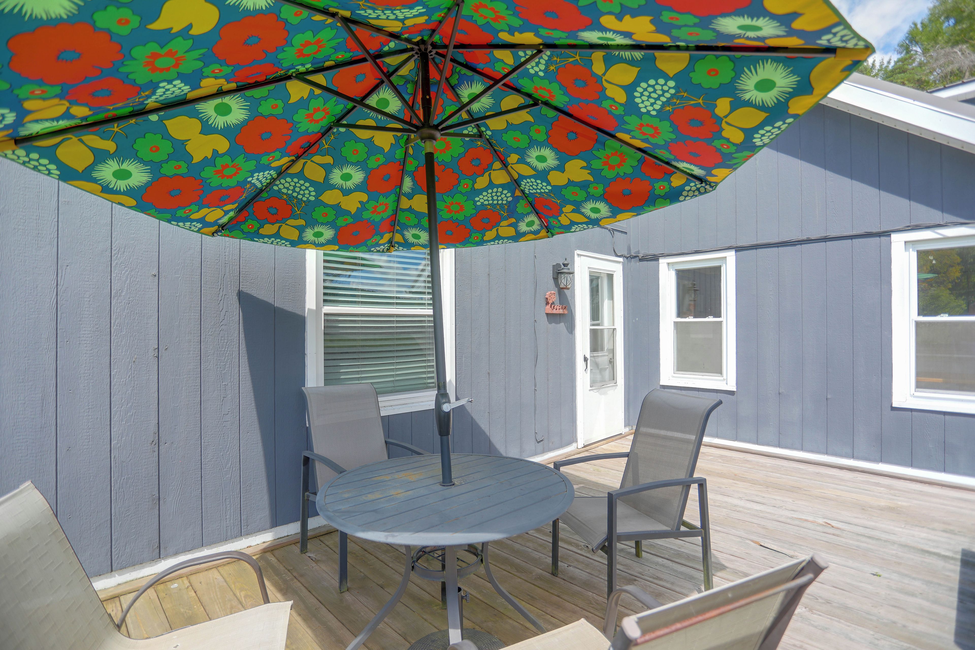 An outdoor patio with a table and chairs, a colorful umbrella, and a house with a door and windows.