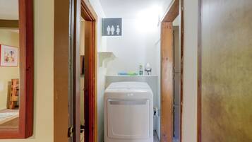A laundry room with a washing machine, a shelf with cleaning supplies, and a door with a sign indicating a men's restroom.