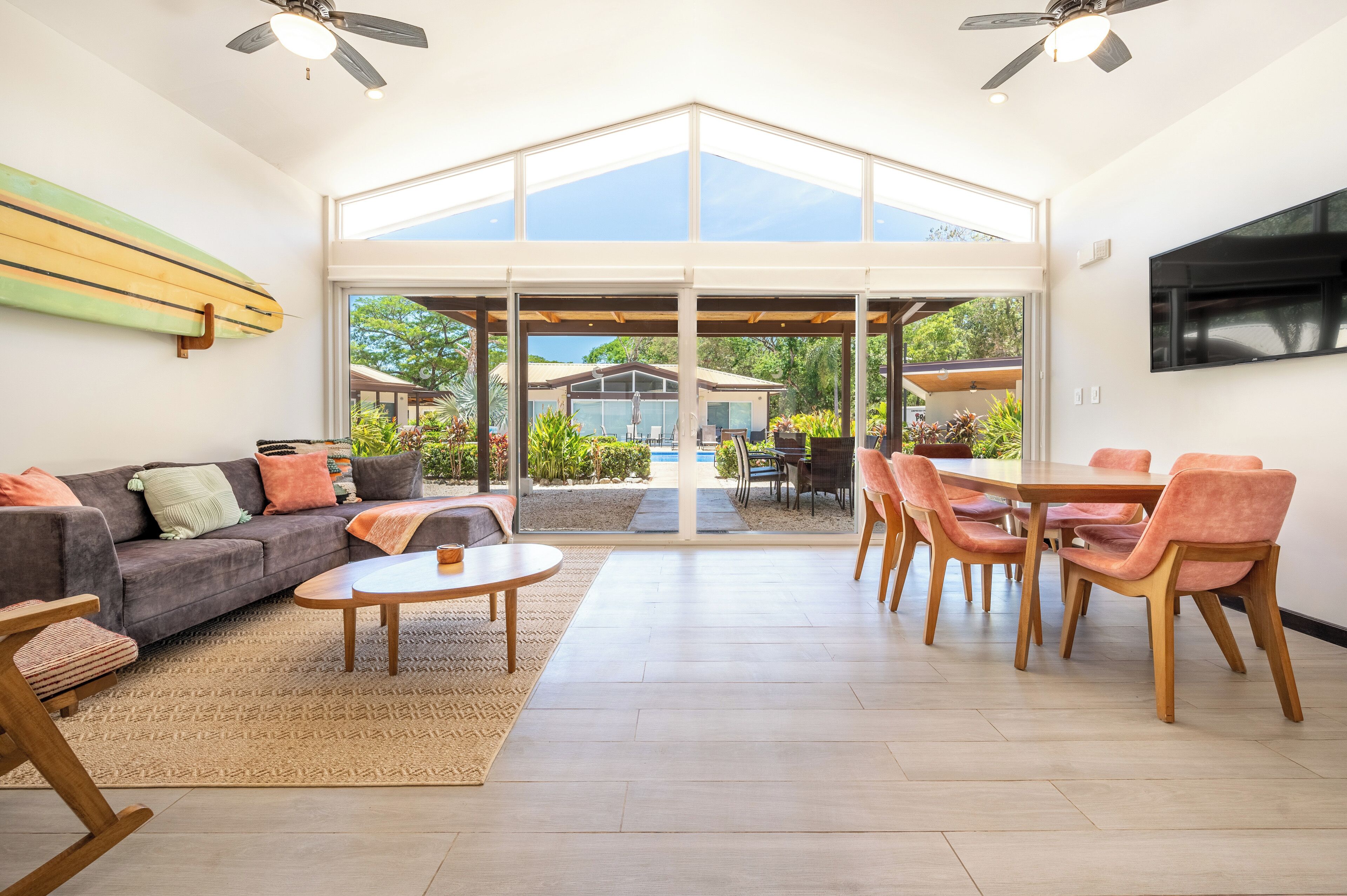 A spacious living area with a large glass door leading to an outdoor patio, a grey sofa, a round wooden table, and a dining set with wooden chairs.