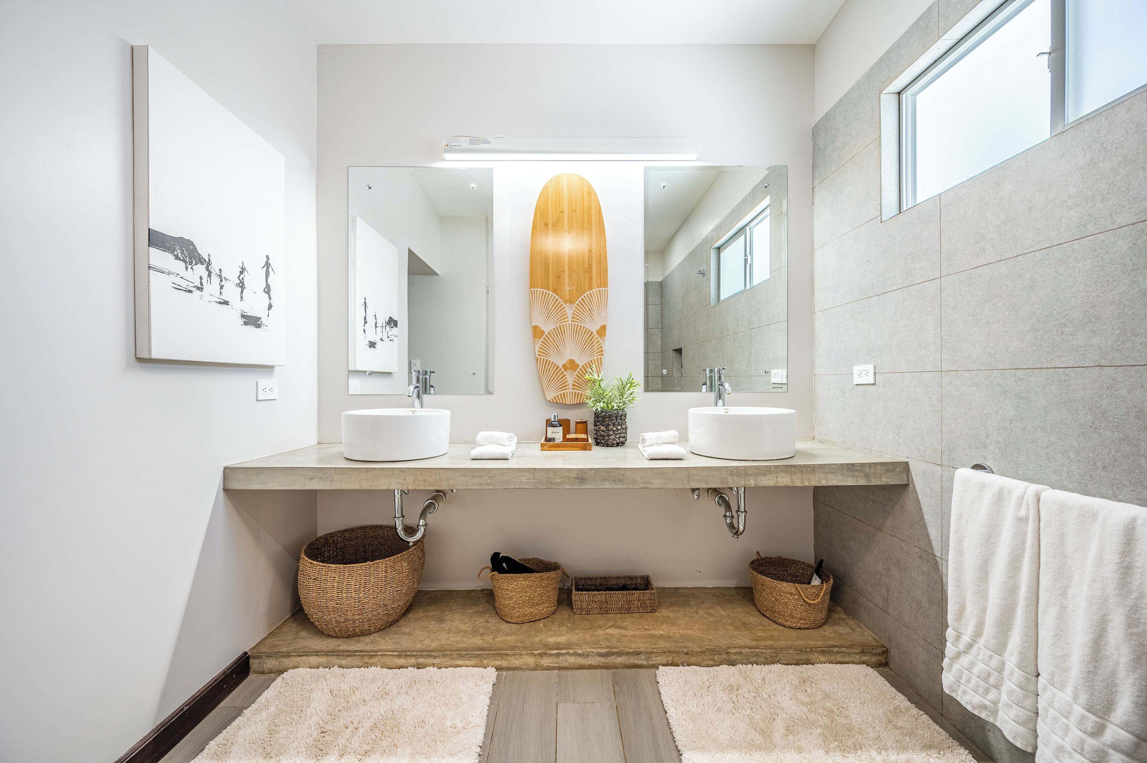 A modern bathroom with two sinks, a large mirror, and a wooden decorative piece above the mirror. There are two woven baskets under the sinks and a white towel hanging on the wall.