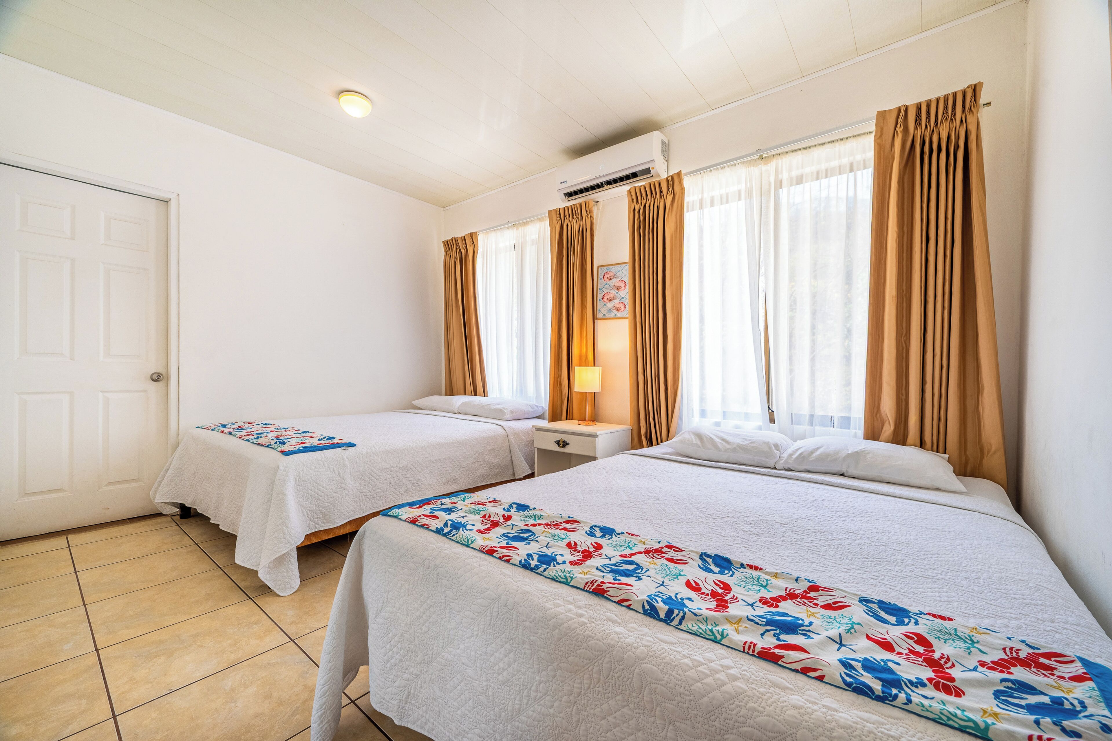 Une chambre d’hôtel avec deux lits, une fenêtre avec des rideaux, une porte et un sol carrelé.