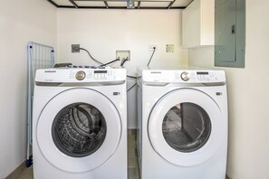 Dos lavadoras Samsung blancas en un lavadero con un tendedero metálico y un enchufe eléctrico en la pared.