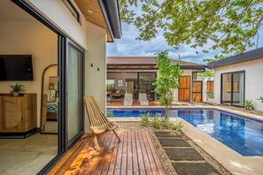 Un área de piscina moderna al aire libre con una terraza de madera, un pequeño árbol y vistas al espacio interior.