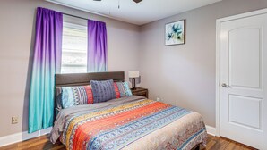 Quarto com cama, mesa de cabeceira, ventoinha no teto, janela com cortinas e porta.