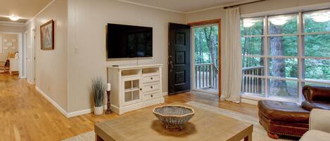 A living room with a wooden coffee table, a brown leather sofa, a television mounted on the wall, and a large window with a view of trees.