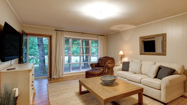 Uma sala de estar com mesa de centro de madeira, poltrona de couro, sofá branco, televisão fixada na parede e uma grande janela com cortinas.
