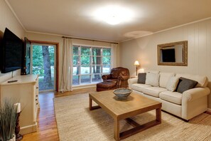 A living room with a wooden coffee table, a leather armchair, a white sofa, a television mounted on the wall, and a large window with curtains. - Basecamp in Skyline, 10 Mi to Walls of Jericho (Scottsboro)