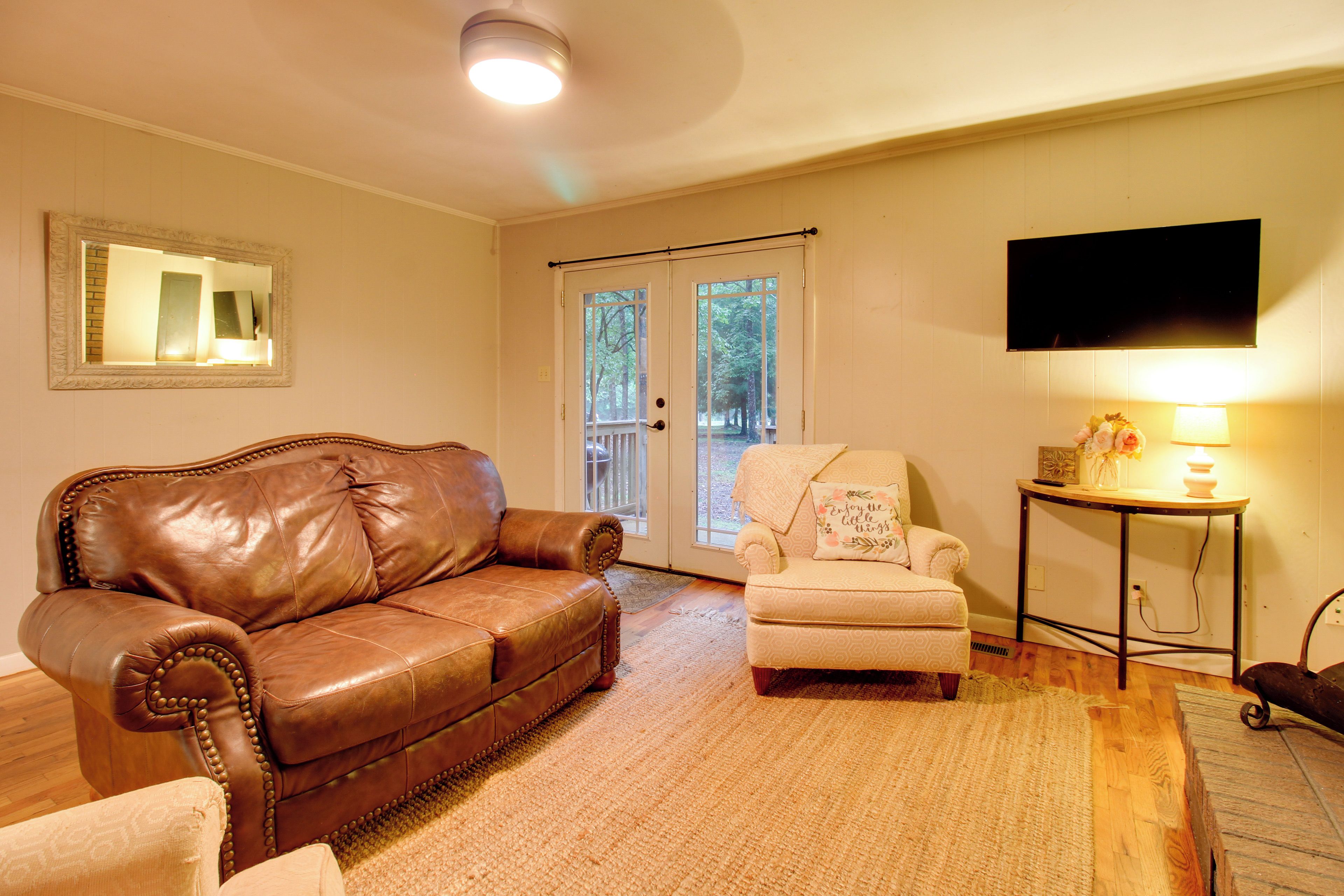 A living room with a brown leather sofa, a beige armchair, a small table with a lamp, and a television on the wall.
