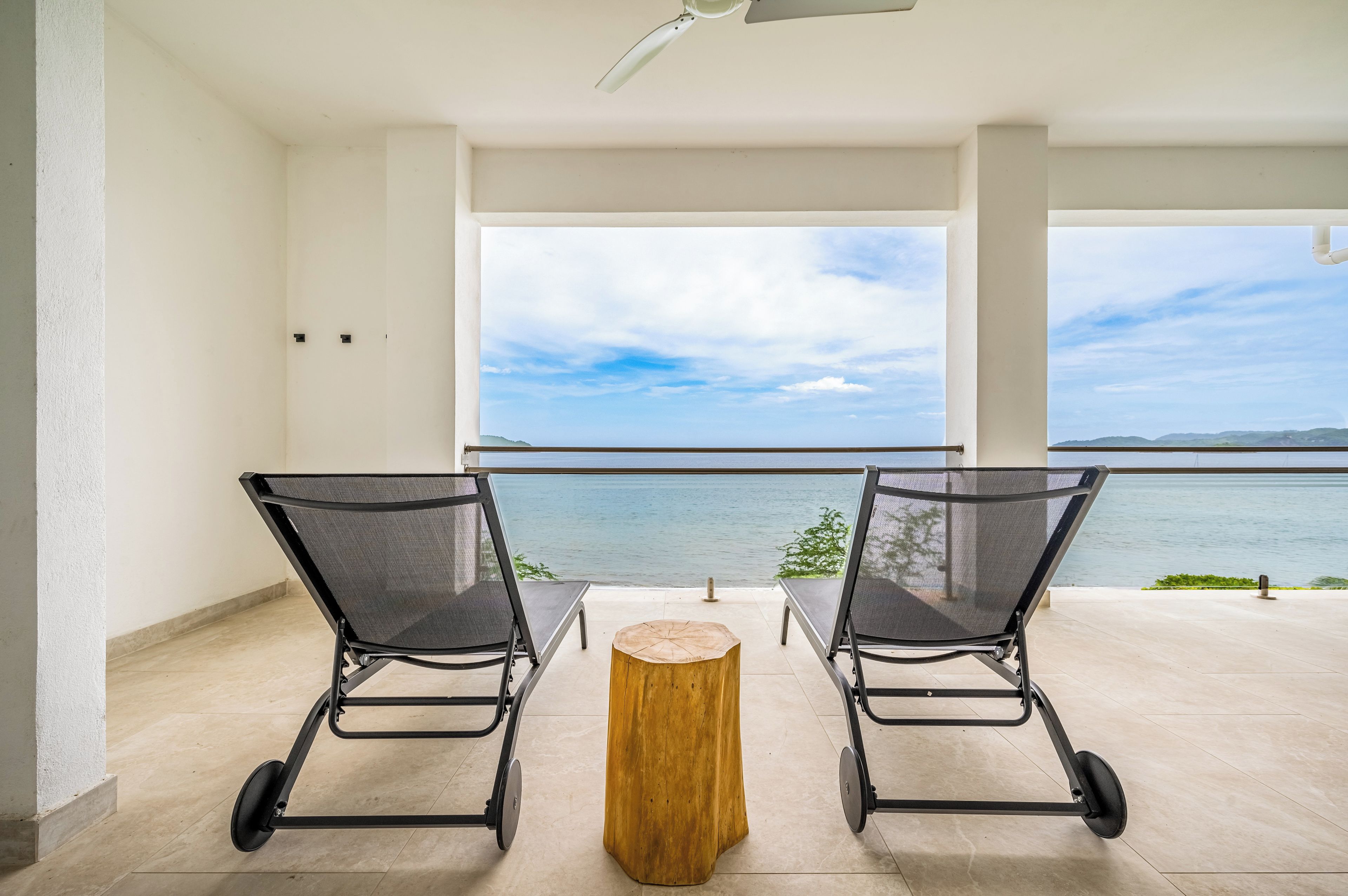 A balcony with two chairs and a wooden stool, overlooking a body of water and distant land.