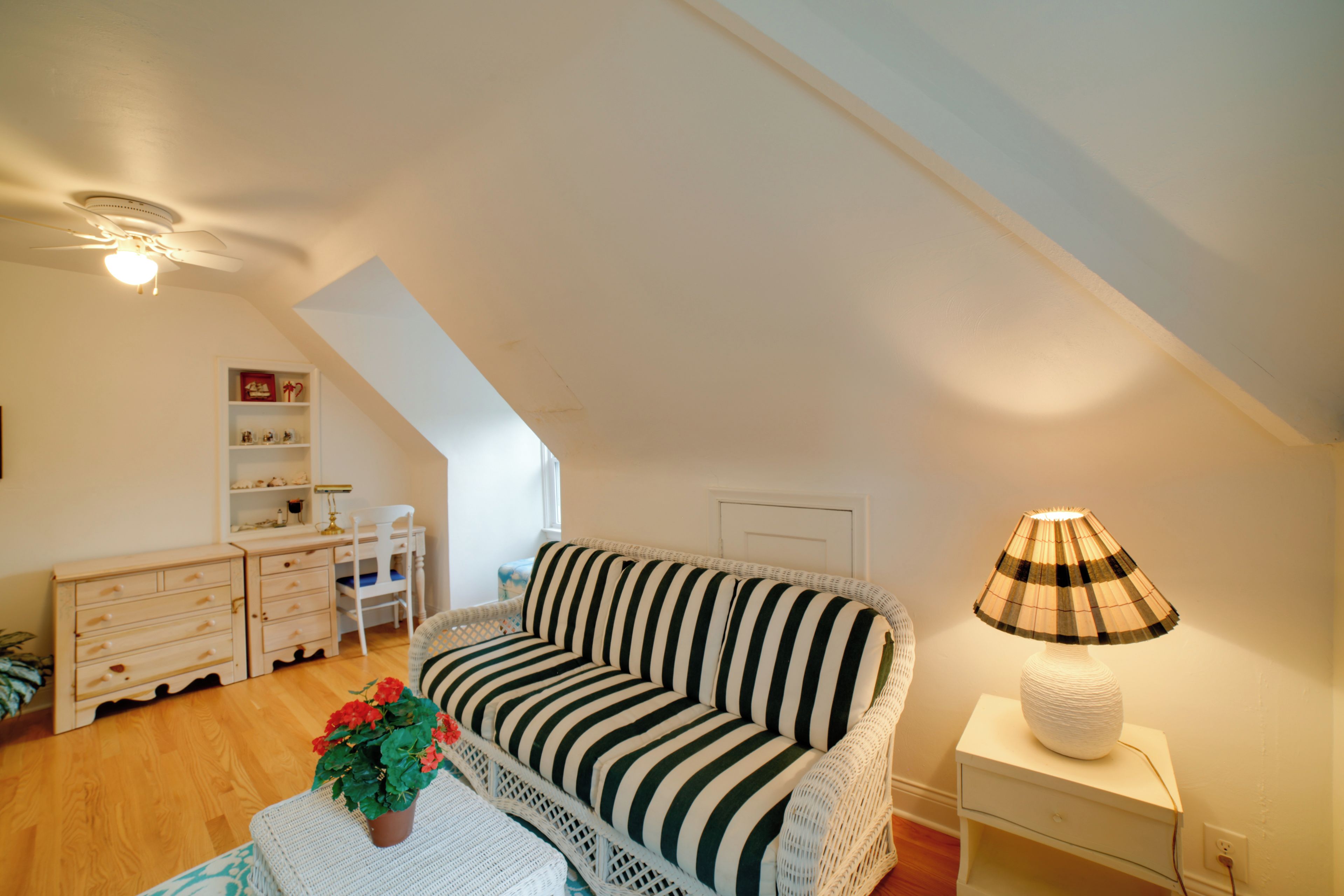 A cozy loft bedroom with a sloped ceiling, a striped sofa, a small table with a plant, a lamp, and a desk with shelves.