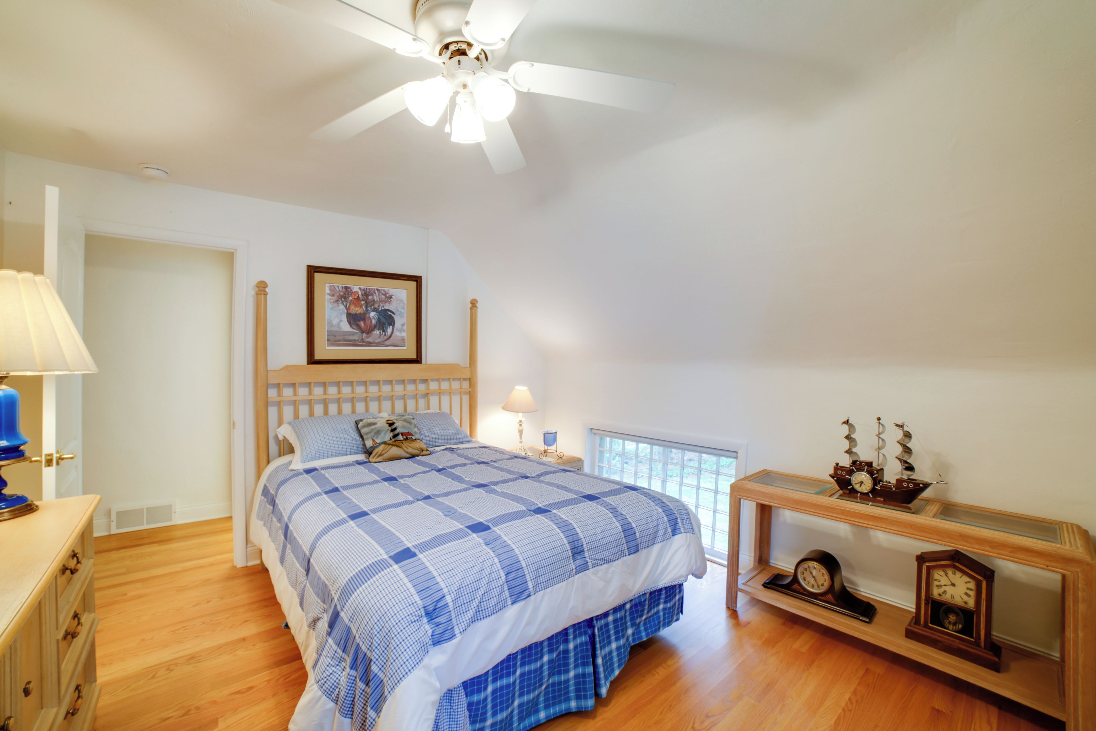 A bedroom with a bed, a ceiling fan, a nightstand, a lamp, a framed picture, and a clock on a shelf.