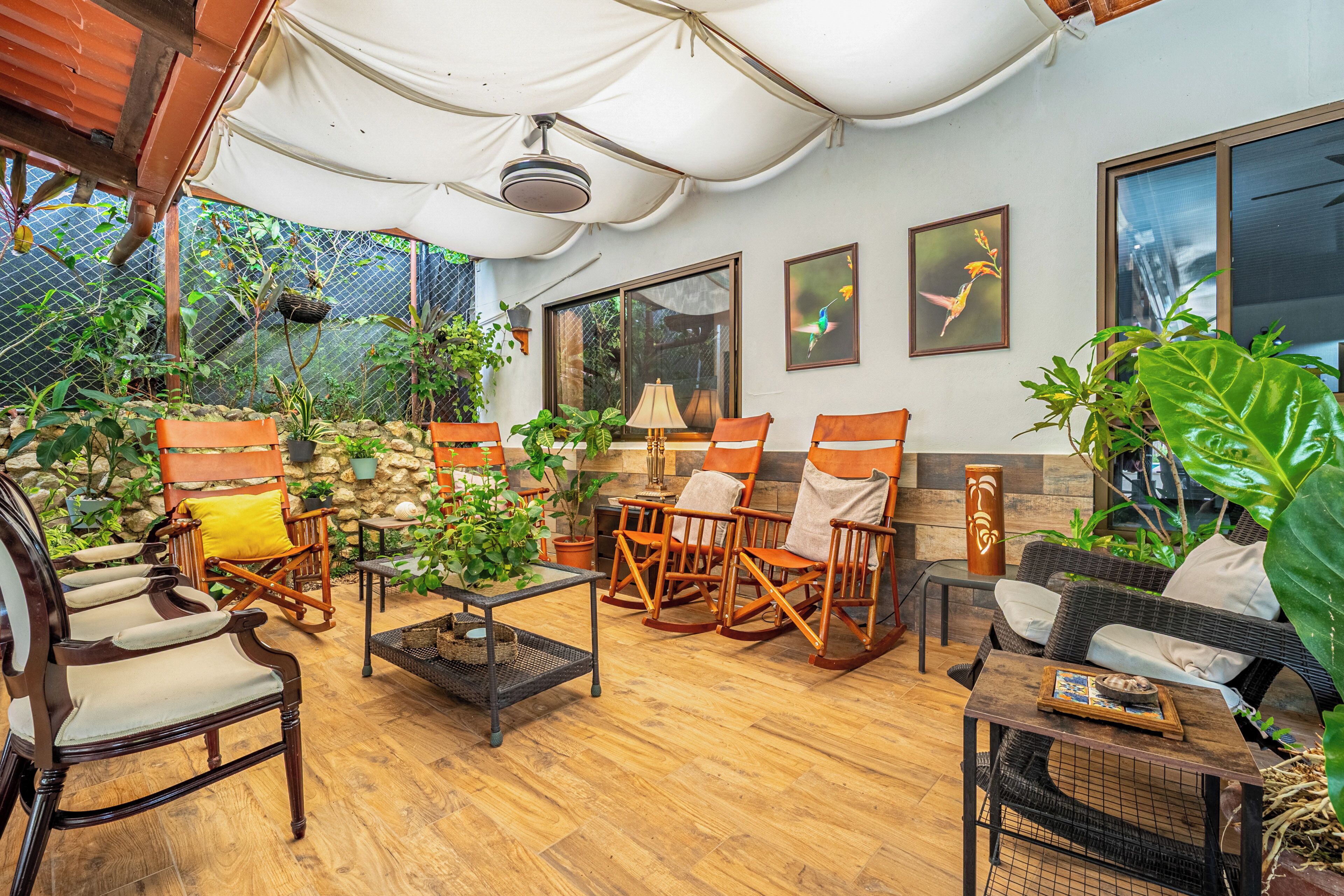 A covered outdoor seating area with wooden furniture, potted plants, and artwork on the walls.