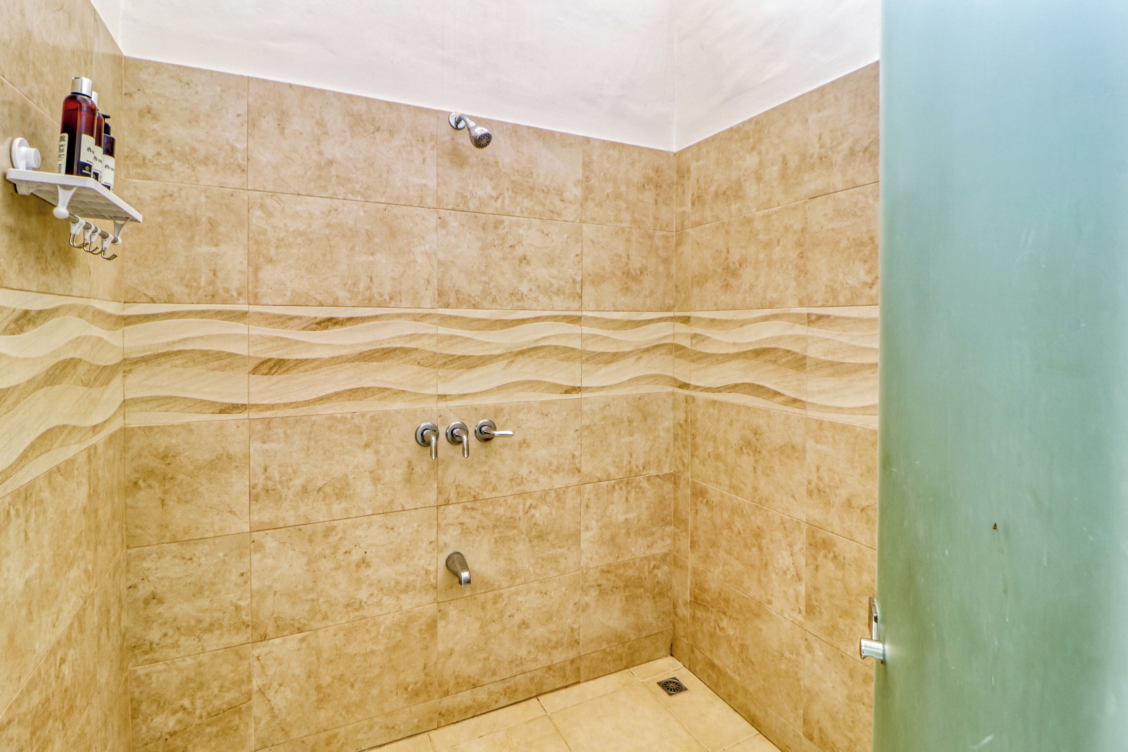 A bathroom with beige tiles, a shower with a handheld showerhead, a wall-mounted faucet, and a shelf with toiletries.