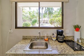 A kitchen with a stainless steel sink, a coffee maker, and a view of a tropical landscape through a window. - Studio w/ Resort-style Pool, Walk to Beach! (Sardinal)