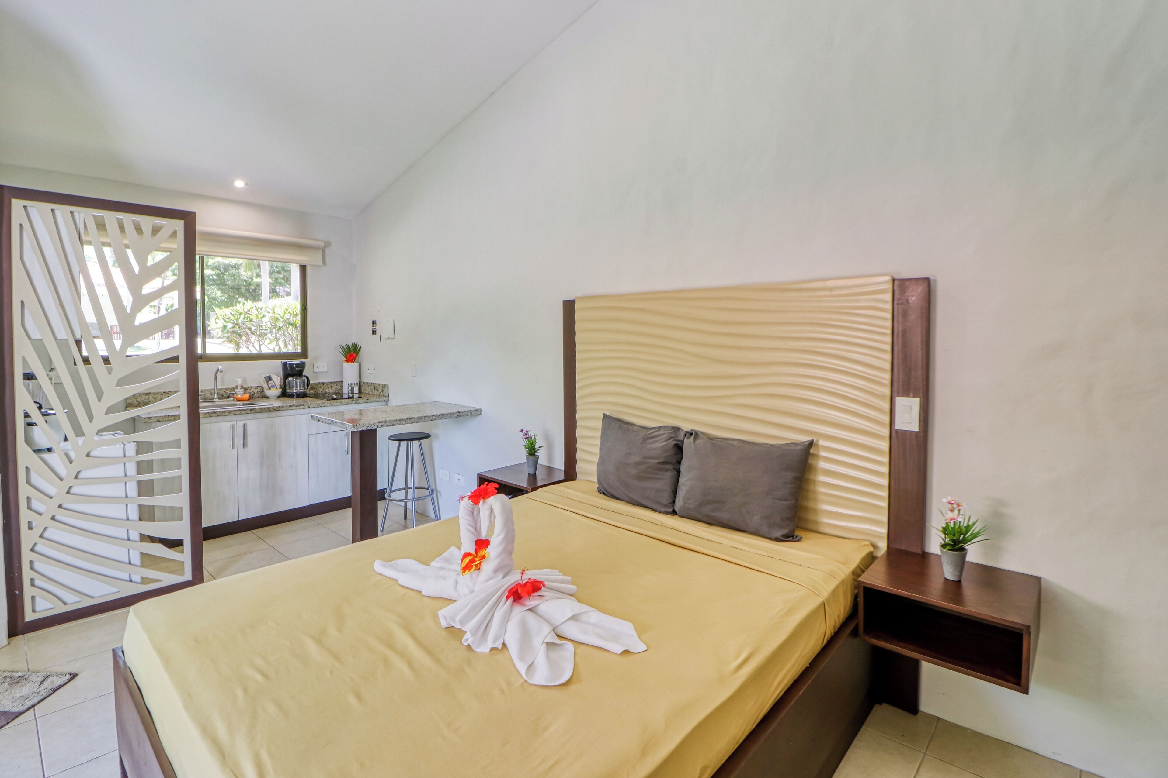 A hotel room with a bed, a nightstand, a small table, a window, and a kitchenette.