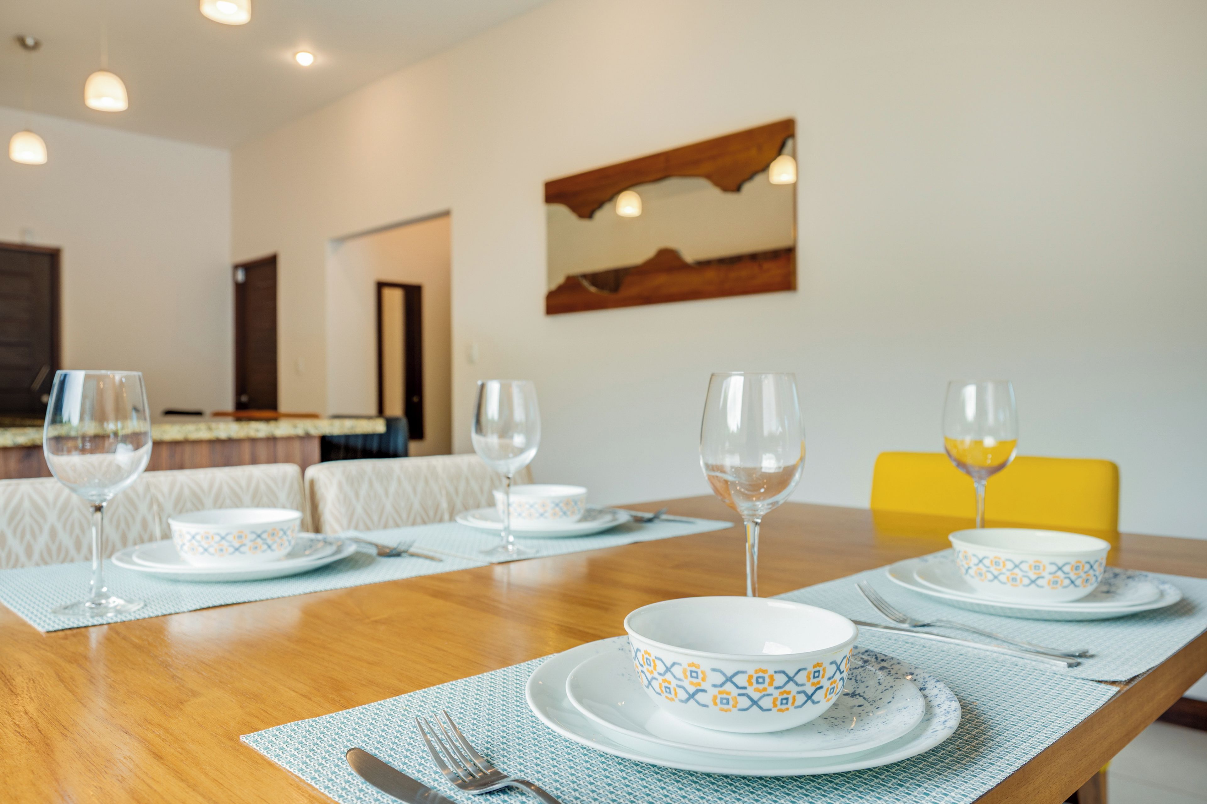 A dining table set with plates, bowls, and wine glasses, with a yellow chair and a painting on the wall.
