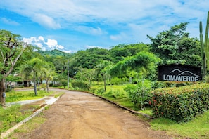 A dirt road leading to a sign with 'LOMAVERDE' written on it, surrounded by lush greenery and trees.