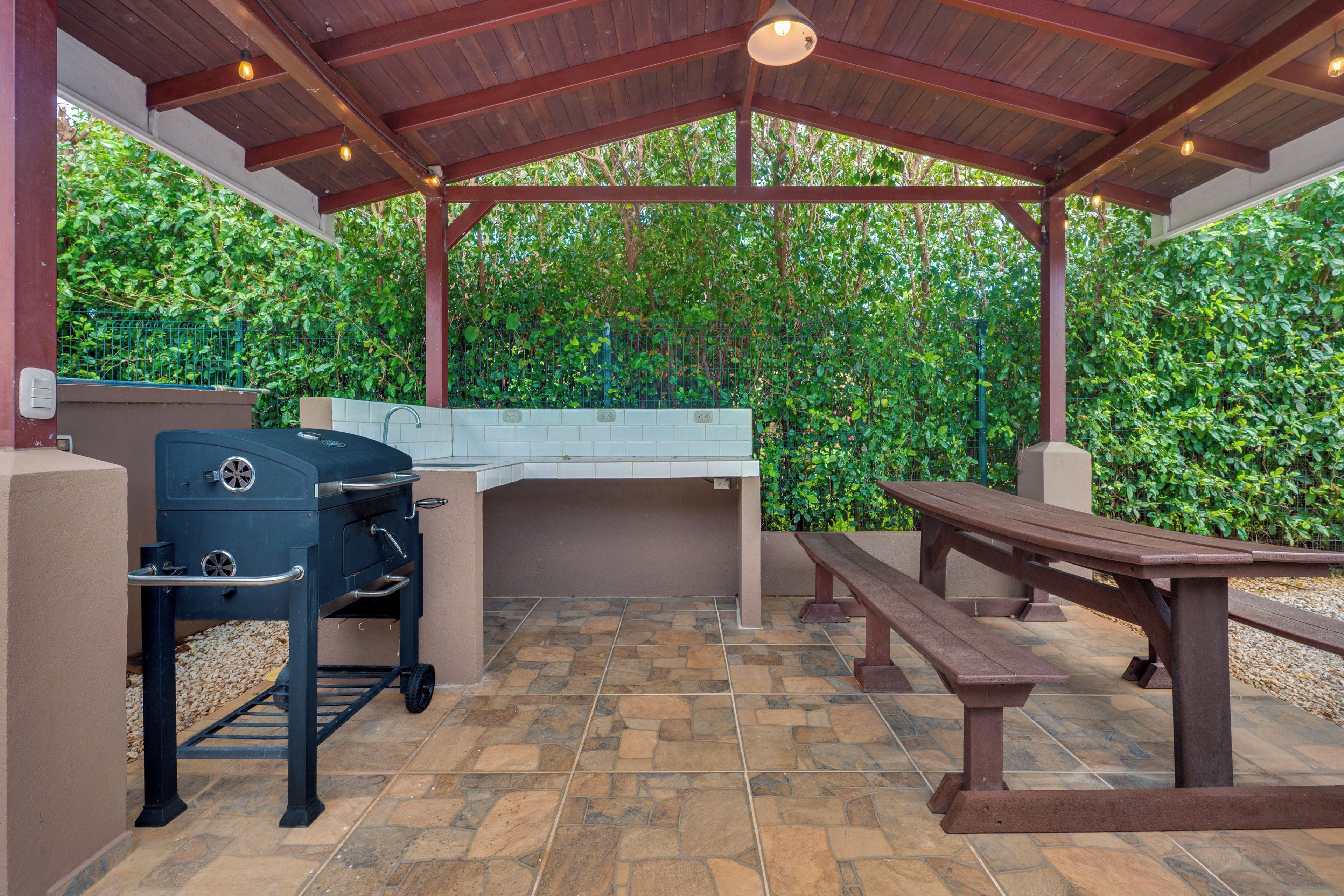 Une terrasse extérieure dotée d’un barbecue, d’un sol carrelé en pierre, d ’un banc en bois et d’un espace couvert avec un toit en métal.