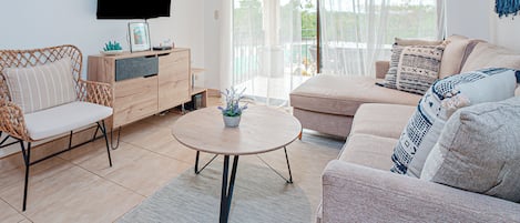 Un salon moderne avec un canapé, des fauteuils, une table basse, un téléviseur et une vue sur l’extérieur, visible à travers une porte vitrée.