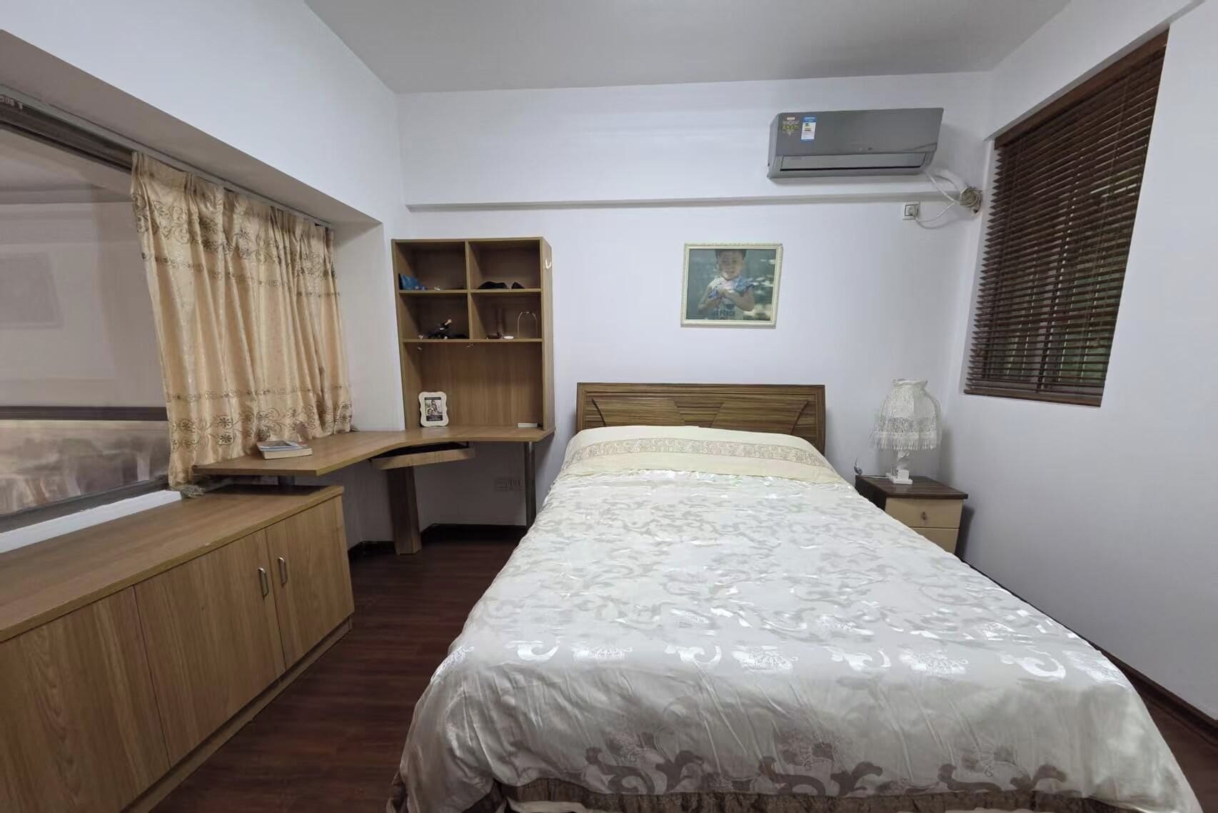 A bedroom with a bed, a desk, a wardrobe, a shelf, a picture on the wall, and an air conditioning unit.