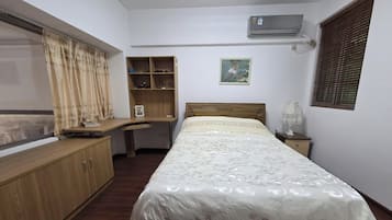 A bedroom with a bed, a desk, a wardrobe, a shelf, a picture on the wall, and an air conditioning unit.