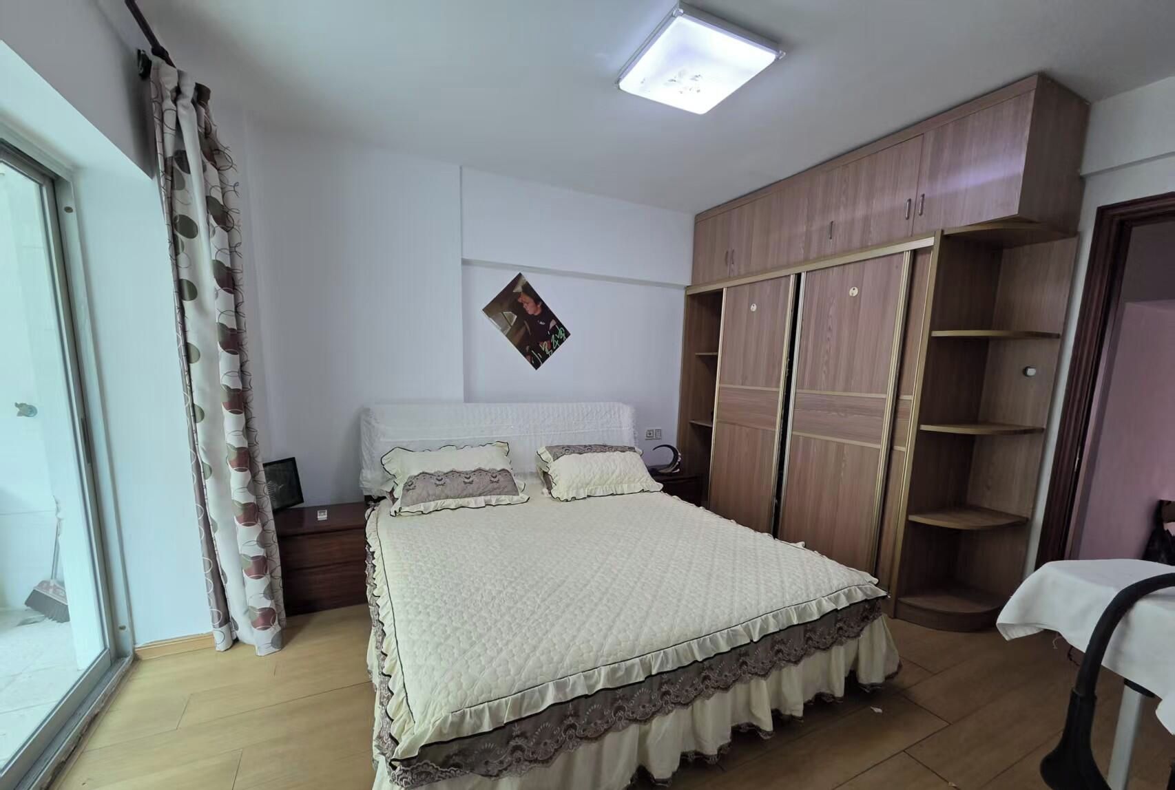 A bedroom with a bed, a wardrobe, a bedside table, and a window with curtains.