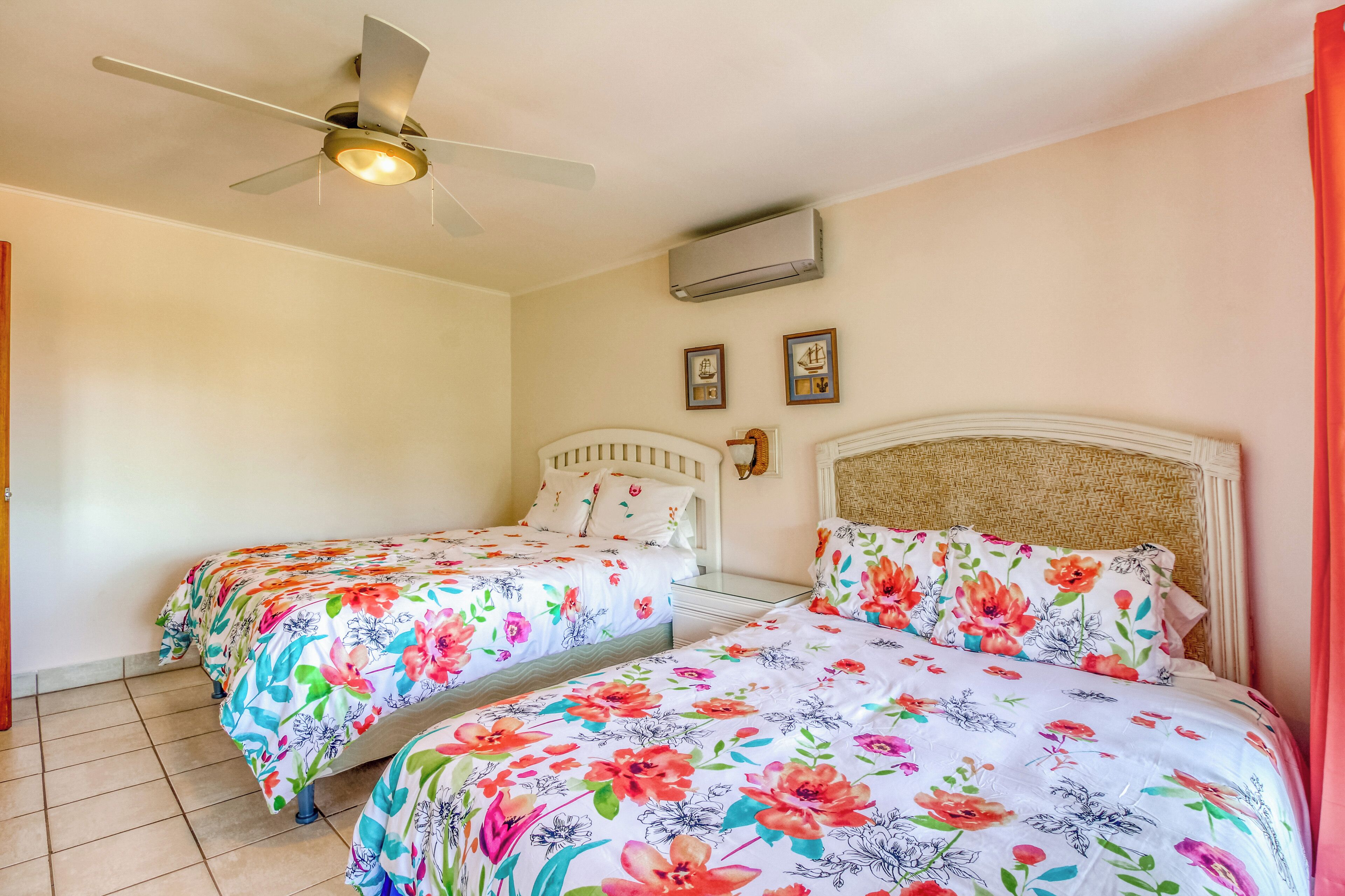 Habitación de hotel con dos camas, un ventilador de techo, un equipo de aire acondicionado y dos cuadros en la pared.