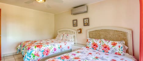 Quarto de hotel com duas camas, um ventilador de teto, ar condicionado e duas fotos emolduradas na parede.