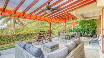 Pátio coberto ao ar livre com móveis de vime, ventiladores de teto e vista para um jardim tropical.
