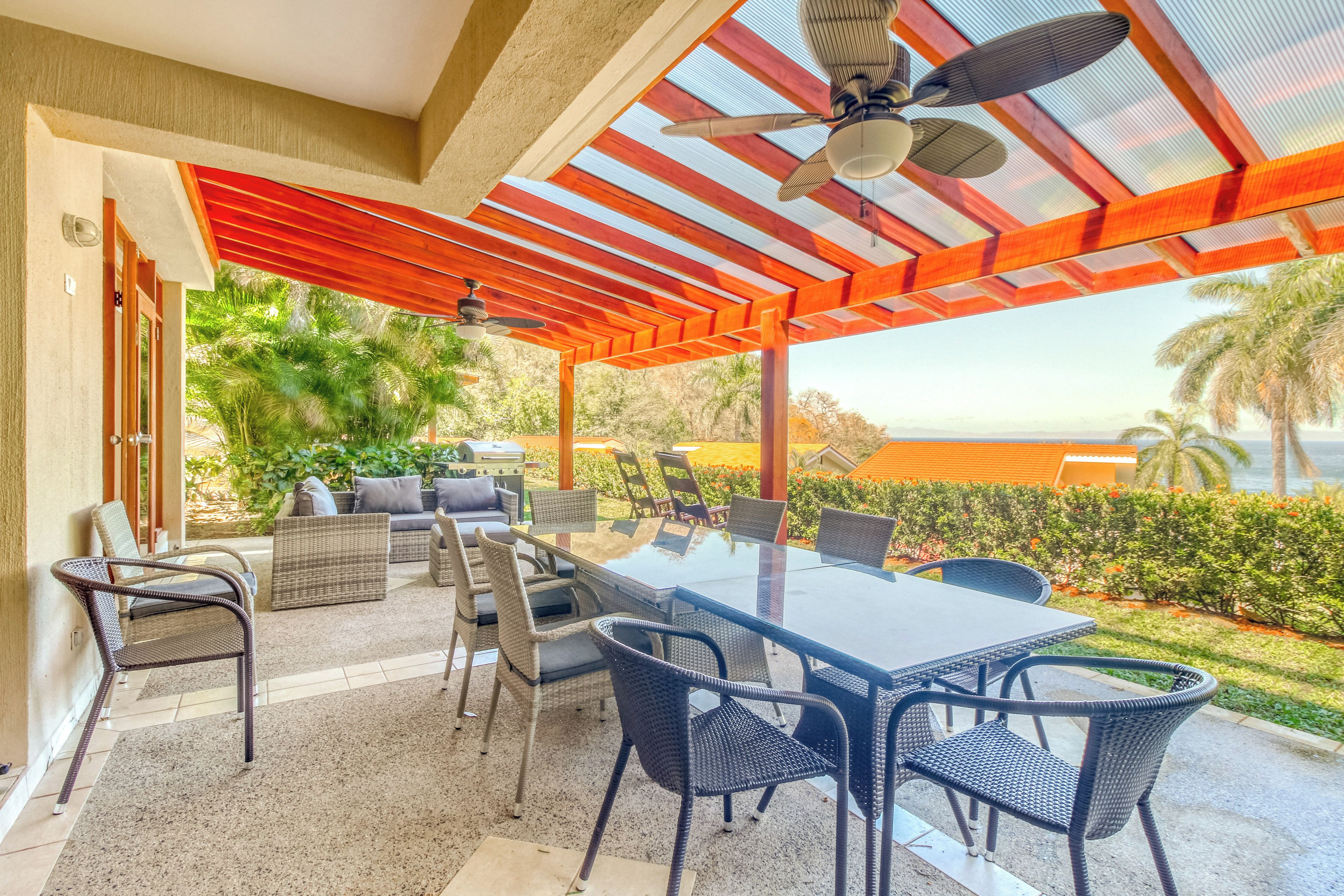 Eine Terrassengruppe im Freien mit einem Glastisch, Stühlen, einem Deckenventilator und Blick aufs Meer.