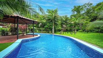 A swimming pool with a tiled edge, surrounded by lush greenery and a covered seating area.