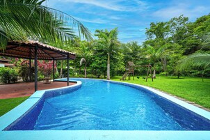 Una piscina con borde de azulejos, rodeada de abundante vegetación y un área de estar techada.