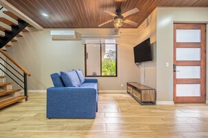 A modern living room with a blue sofa, wooden flooring, a ceiling fan, a TV mounted on the wall, and a staircase with wooden railings.