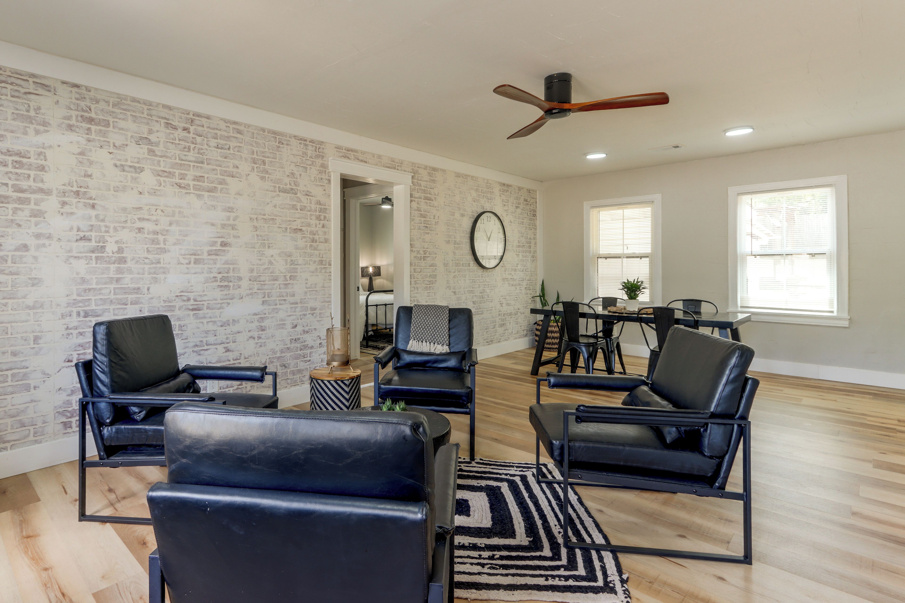 A spacious living room with a brick wall, a ceiling fan, a dining area with a table and chairs, and a large clock on the wall.