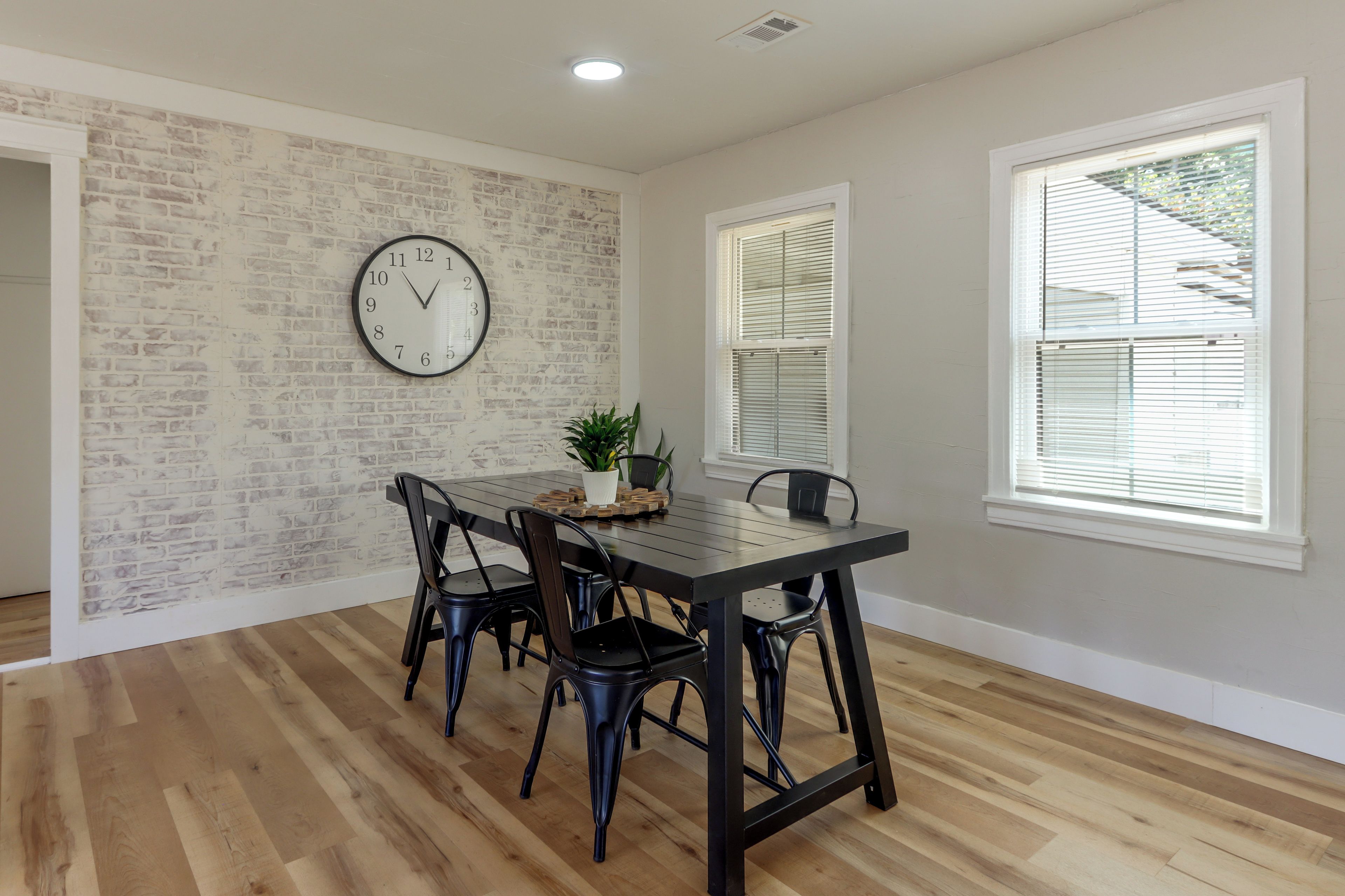 A dining area with a brick wall, a round wall clock, and a window with blinds.