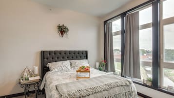 A bedroom with a large bed, a bedside table, a window with curtains, and a wall-mounted plant.